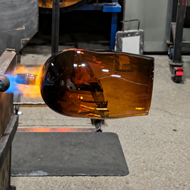 An amber-colored glass vessel, likely a bowl or mold, held in a glassblowing workshop under a torch with a blue flame visible on the left.