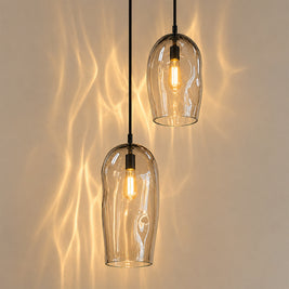Two modern pendant light fixtures with clear glass bell-shaped shades and black cords, hanging against a beige wall with warm light cast across it.